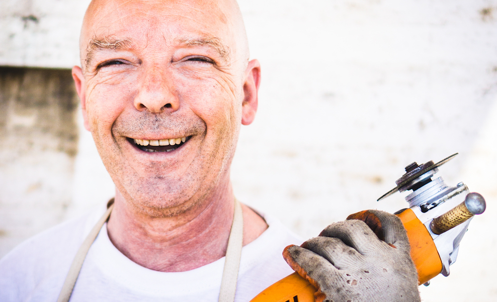 Older man smiles brightly while holding a tool.